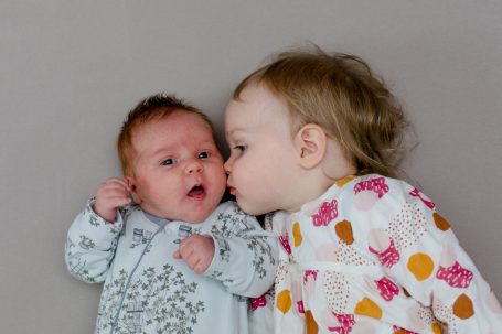 Ein Kleinkind küsst ein Baby auf die Wange, beide liegen auf einem grauen Untergrund.