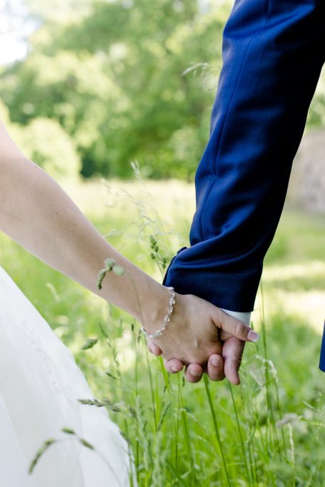 Hochzeitskleidung, Hände eines Paares halten sich in einer grünen Wiese.