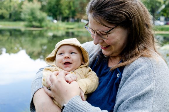 Eine lächelnde Frau hält ein Baby am Seeufer. Beide tragen fröhliche Kleidung.