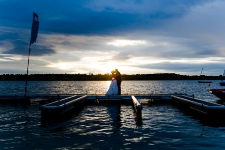 Hochzeitspaar steht am Ufer bei Sonnenuntergang, umgeben von Wasser und Wolken.