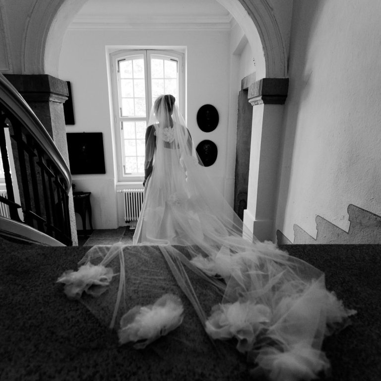 Braut in einem weißen Kleid mit Schleier, stehend auf einer Treppe in einem hellen Raum.