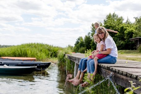 Zwei Kinder spielen am Ufer, während eine Frau sie umarmt, Boote sind im Hintergrund.