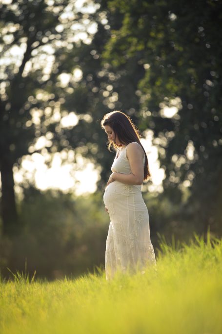 Schwangere Frau in einem weißen Kleid steht in einem grünen Feld.