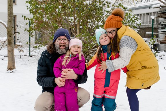 Familie mit zwei Mädchen im Schnee, lächelnd und umarmend, in winterlicher Kleidung.
