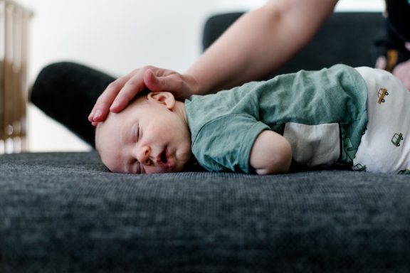 Schlafender Säugling auf einer Couch, sanft von einer Hand gestreichelt.