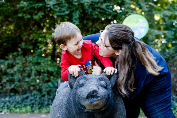 Ein Kind sitzt auf einem Bären-Skulptur und lacht, während eine Frau ihm einen Kuss gibt.