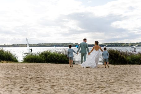 Brautpaar mit zwei Kindern am Strand, Blick auf den See und Segelboote im Hintergrund.