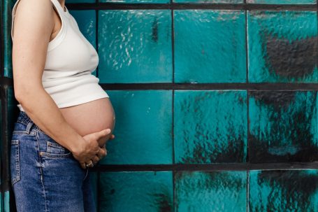 Schwangere Person in einem weißen Top, die ihren Bauch vor einer türkisfarbenen Wand hält.