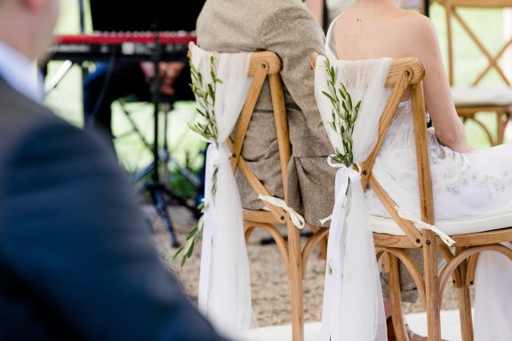 Stühle bei einer Hochzeit, dekoriert mit Pflanzen und Bändern. Im Hintergrund Musiker.