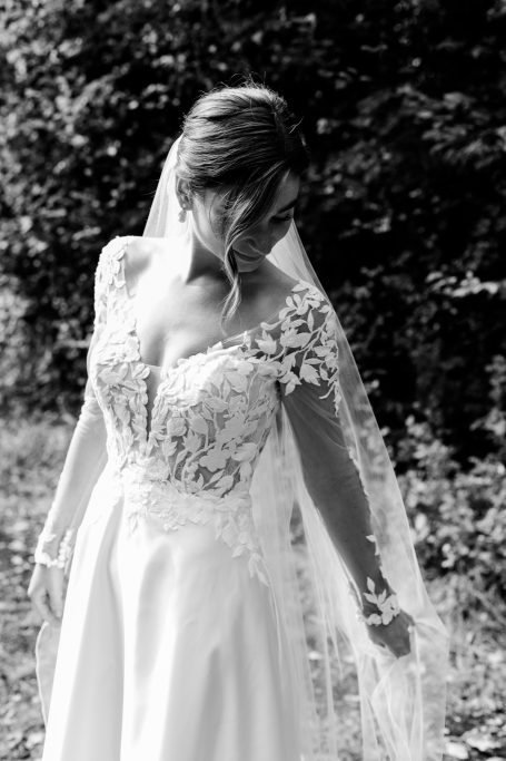 Braut in elegantem Kleid mit Spitze und Schleier, vor einer grünen Kulisse.