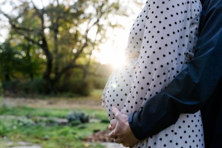Ein schwangerer Bauch, umarmt von einer Person, mit Sonnenstrahlen im Hintergrund.