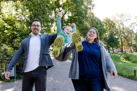 Ein glückliches Paar hebt ein Kind im Park mit grünen Schuhen in die Luft.