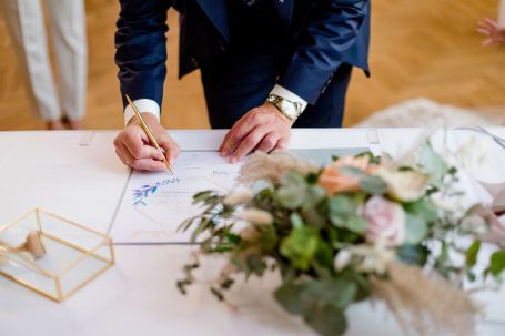 Eine Hand schreibt auf ein Dokument, umgeben von Blumen und einer goldenen Box.