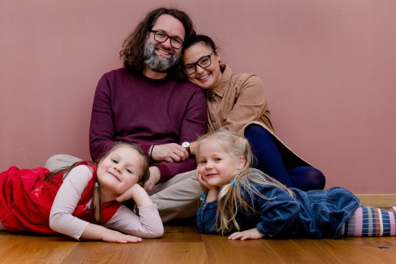 Familie sitzt auf dem Boden, zwei Mädchen und zwei Erwachsene lächeln fröhlich.