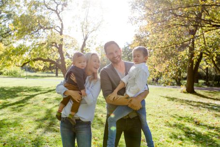 Familie mit zwei Kindern in einem Park, umgeben von Bäumen und Sonnenlicht.