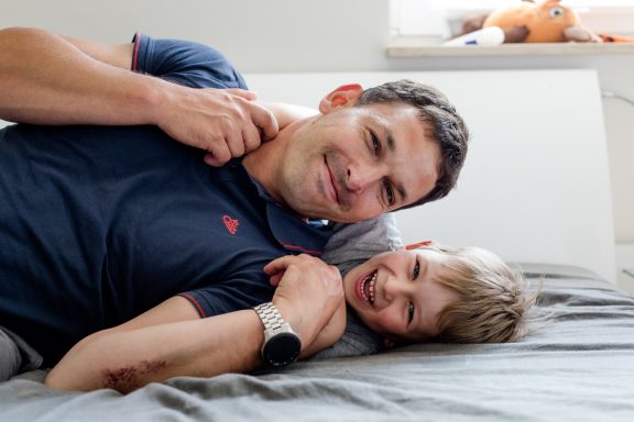 Vater und Sohn liegen lächelnd auf dem Bett und haben Spaß miteinander.