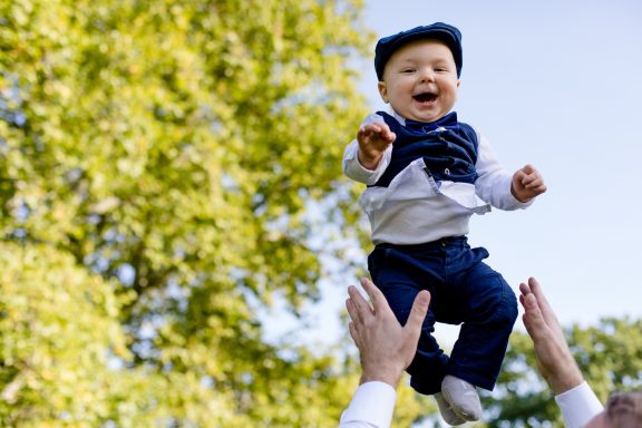 Ein fröhliches Baby wird von Händen in die Luft gehoben, umgeben von Bäumen.