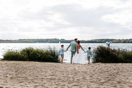 Familie mit zwei Kindern am Strand, Hand in Hand, vor einem See mit sanften Wellen.