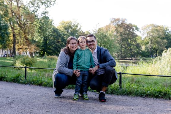 Familie posiert im Park: Mutter, Sohn und Vater lächeln zusammen vor einer grünen Landschaft.