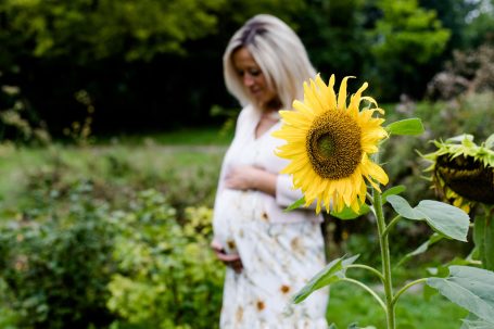 Schwangere Frau in einem weißen Kleid steht neben einer Sonnenblume im Freien.