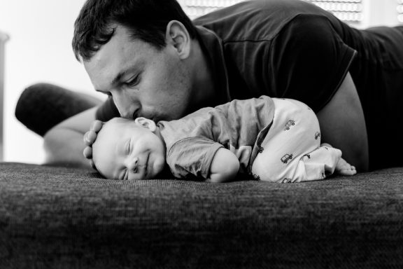 Vater küsst lächelnd sein schlafendes Baby auf einem Sofa.