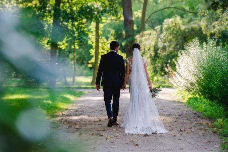 Braut und Bräutigam gehen Hand in Hand einen Weg im Garten entlang.