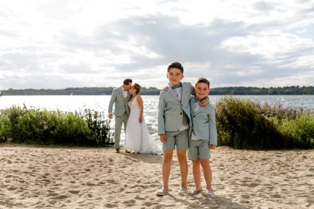 Zwei Jungen in Anzügen stehen am Strand, im Hintergrund ein Hochzeits-Paar.