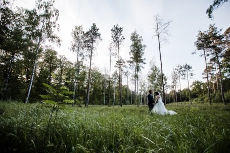 Ein Paar steht in einem Wald, umgeben von hohen Bäumen und grüner Wiese.
