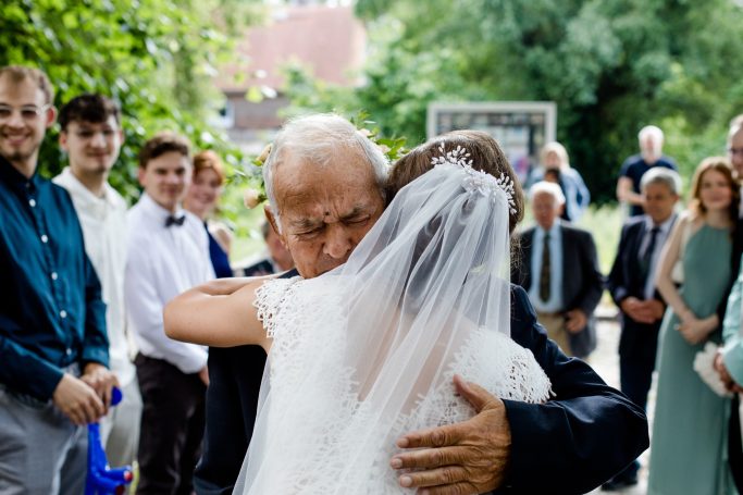 Ein älterer Mann umarmt eine Braut in einem festlichen Moment, umgeben von Gästen.