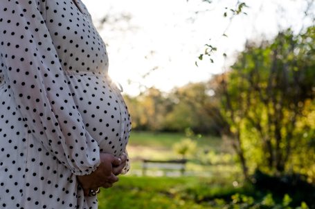 Schwangere Frau hält ihren Bauch, umgeben von einer grünen Landschaft und sanftem Licht.