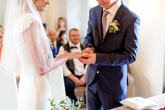 Ein Brautpaar steckt sich während der Zeremonie die Ringe an. Gäste im Hintergrund.