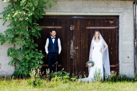 Braut in weißem Kleid mit Blumenstrauß und Bräutigam in Anzug vor einer Holztür.