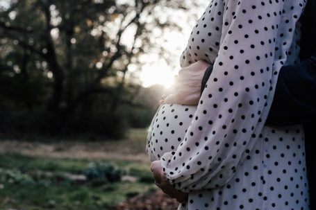 Schwangere Person in gepunktetem Kleid, von hinten umarmt, mit Sonnenlicht im Hintergrund.