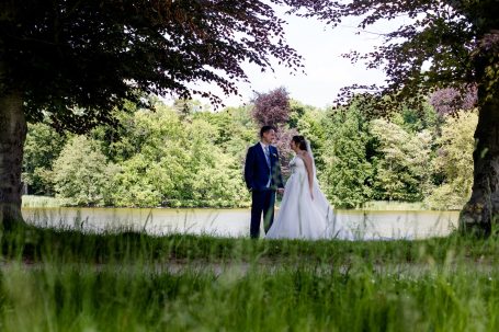 Paar in Hochzeitskleidung, umgeben von Bäumen und Wiese, am Ufer eines Sees.