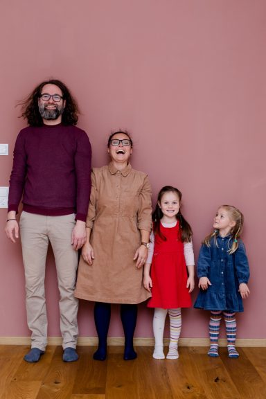 Vier Personen stehen vor einer rosa Wand: zwei Erwachsene und zwei Kinder, die unterschiedlich gekleidet sind.