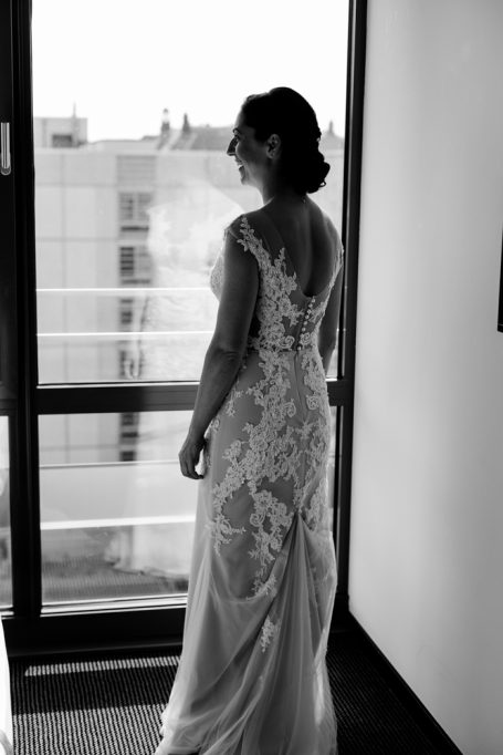 Braut in elegantem Kleid steht vor Fenster und schaut nach draußen.