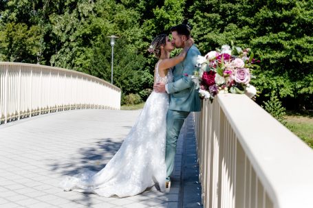 Braut und Bräutigam umarmen sich auf einer Brücke, umgeben von bunten Blumen und Natur.