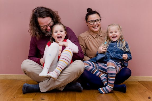 Familie sitzt lächelnd auf dem Boden vor einer rosa Wand, zwei Kinder mit bunten Strümpfen.