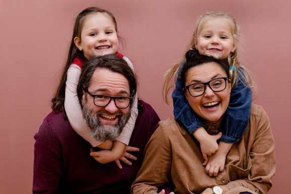 Familie mit zwei kleinen Mädchen, die auf den Schultern der Erwachsenen sitzen und lachen.