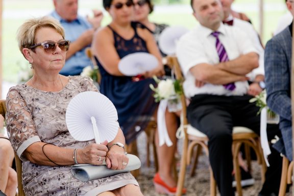 Eine Frau im Vordergrund hält einen Fächer, während Gäste einer Zeremonie sitzen.