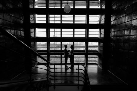 Zwei Silhouetten stehen an einer Treppe vor großen Fenstern in einem modernen Gebäude.