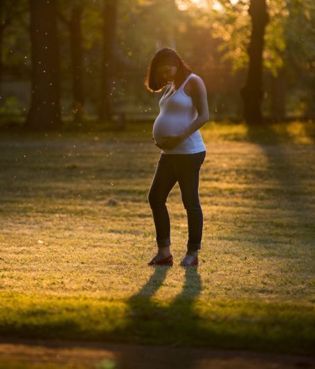 Schwangere Frau mit sichtbarem Bauch steht in einem sonnigen Park.