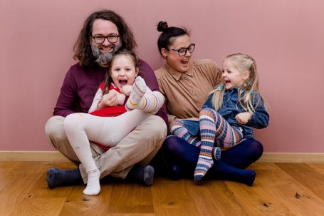 Familie mit zwei Kindern, die fröhlich auf dem Boden vor einer rosa Wand sitzt.