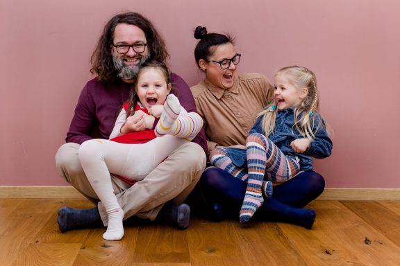 Eine Familie mit zwei Erwachsenen und zwei Kindern lächelt fröhlich vor einer rosa Wand.