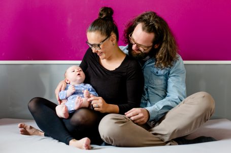 Ein Elternpaar sitzt mit ihrem Baby auf dem Boden vor einer pinken Wand.