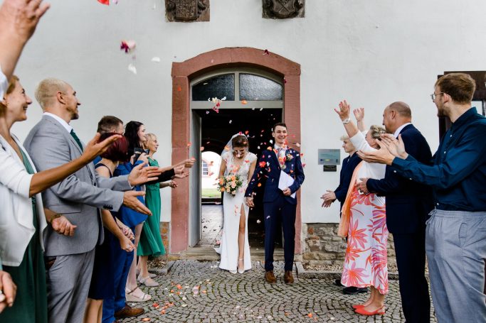 Brautpaar verlässt die Kapelle, umgeben von applaudierenden Gästen und Blumenblüten.