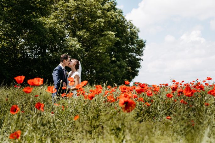 Paar in einem Feld voller roter Mohnblumen, umgeben von grünen Bäumen und blauem Himmel.