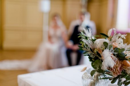 Hochzeitsarrangement mit Blumen im Vordergrund, Brautpaar unscharf im Hintergrund.