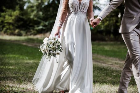 Braut in eleganter weißer Robe hält Blumenstrauß und Hand des Bräutigams.