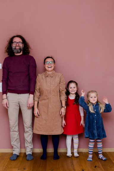 Eine Familie aus vier Personen steht vor einer rosa Wand, lächeln und posieren.